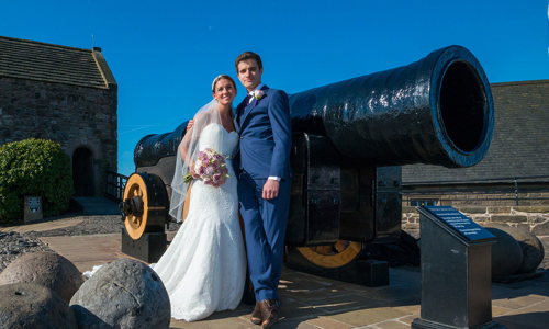 Bride and groom standing beside Mons Meg with St Margaret's Chapel in the background