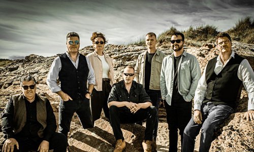 Skipinnish members crouching on rocks, most wearing sunglasses, with sand and grass in the background