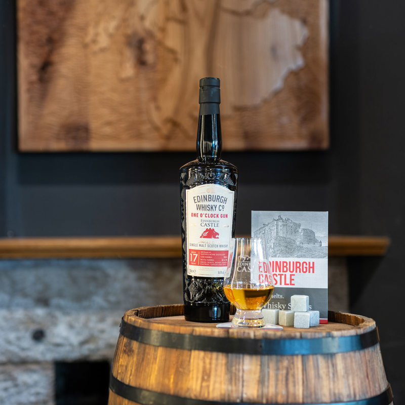 A bottle of One O'Clock Gun Whisky on a barrel with an Edinburgh Castle Whisky glass and stone ice cubes