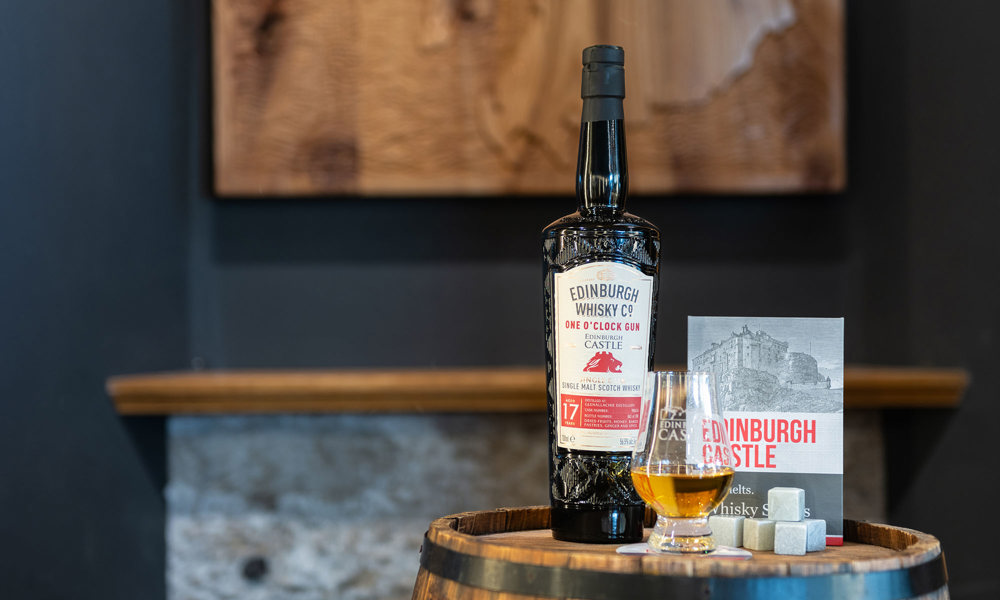 A bottle of One O'Clock Gun Whisky on a barrel with an Edinburgh Castle Whisky glass and stone ice cubes