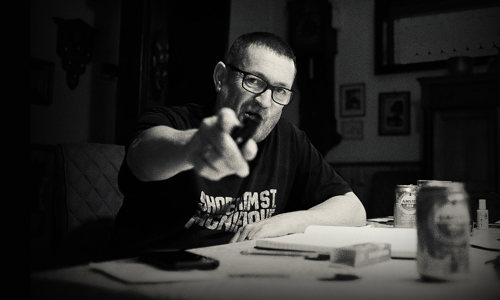Promotional portrait of Paul Heaton sitting at a table pointing at the camera