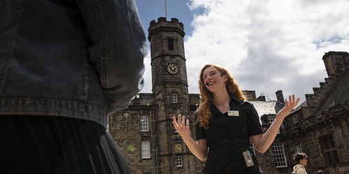 A lady in black and white uniform standing in Crown Square at Edinburgh Castle wearing a portable wireless receiver and headset