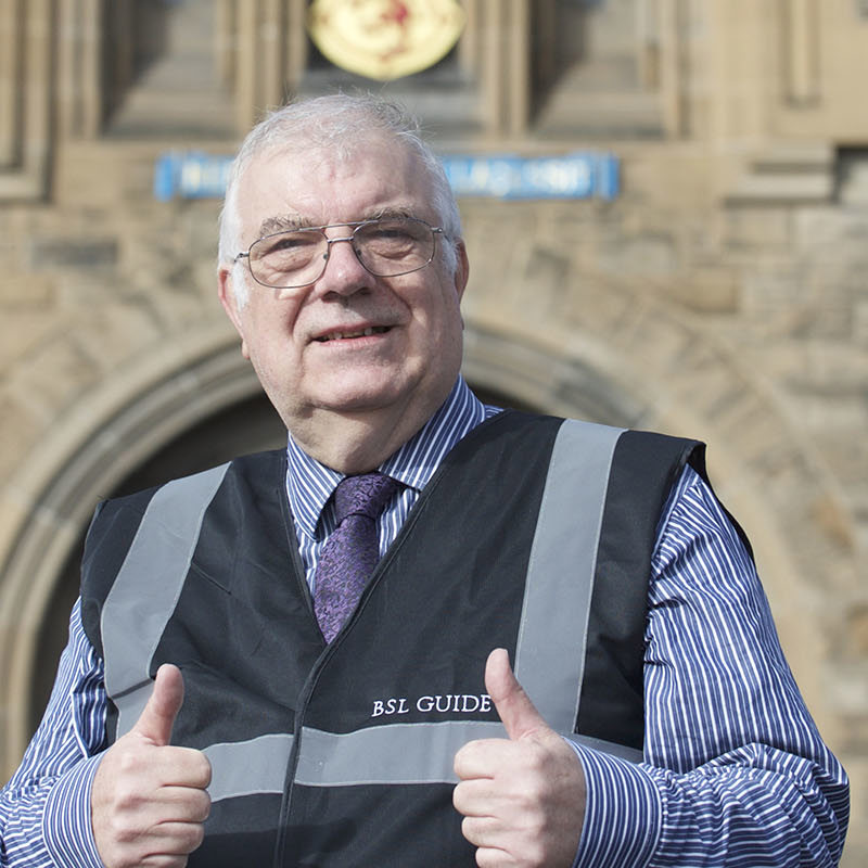 BSL tour guide, John Hay, standing in front of Edinburgh Castle giving two thumbs up