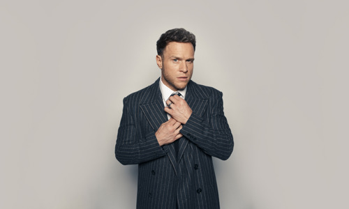 Promotional portrait of Olly Murs in a suit tightening his tie