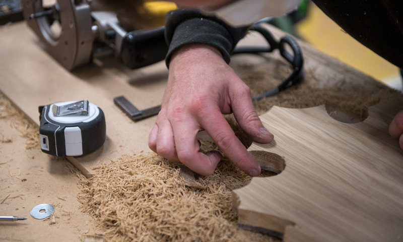 A piece of wood with lots of shavings alongside a persons hand, measuring tape and safety glasses
