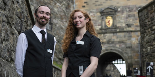 Two people in black and white uniform standing in the entrance to Edinburgh Castle wearing portable wireless receiver and headset