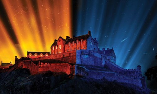 A castle on a rock at night. Half of the castle is illuminated with yellow, and half illuminated with blue, with the colours projecting into the sky.