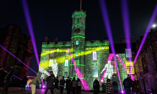 Photo of crown square during Castle of Light. There are projections of Robert the Bruce on the wall and groups of people looking at it.