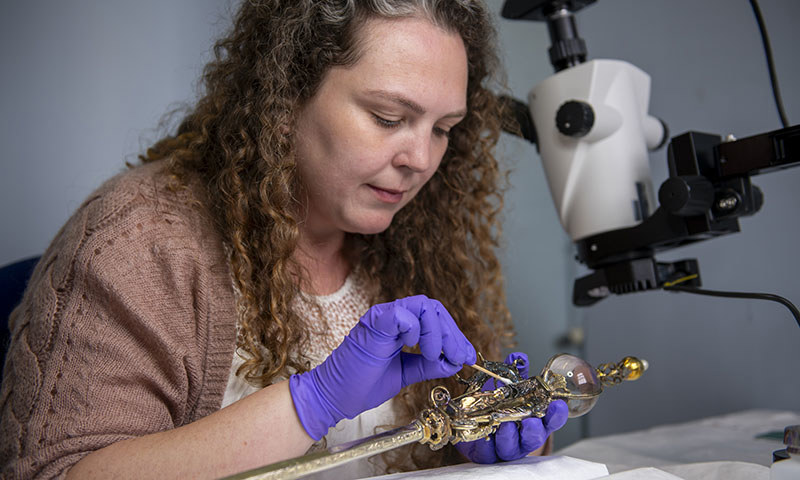 A woman wearing purple gloves holding a Sceptre and looking at it through a microscope